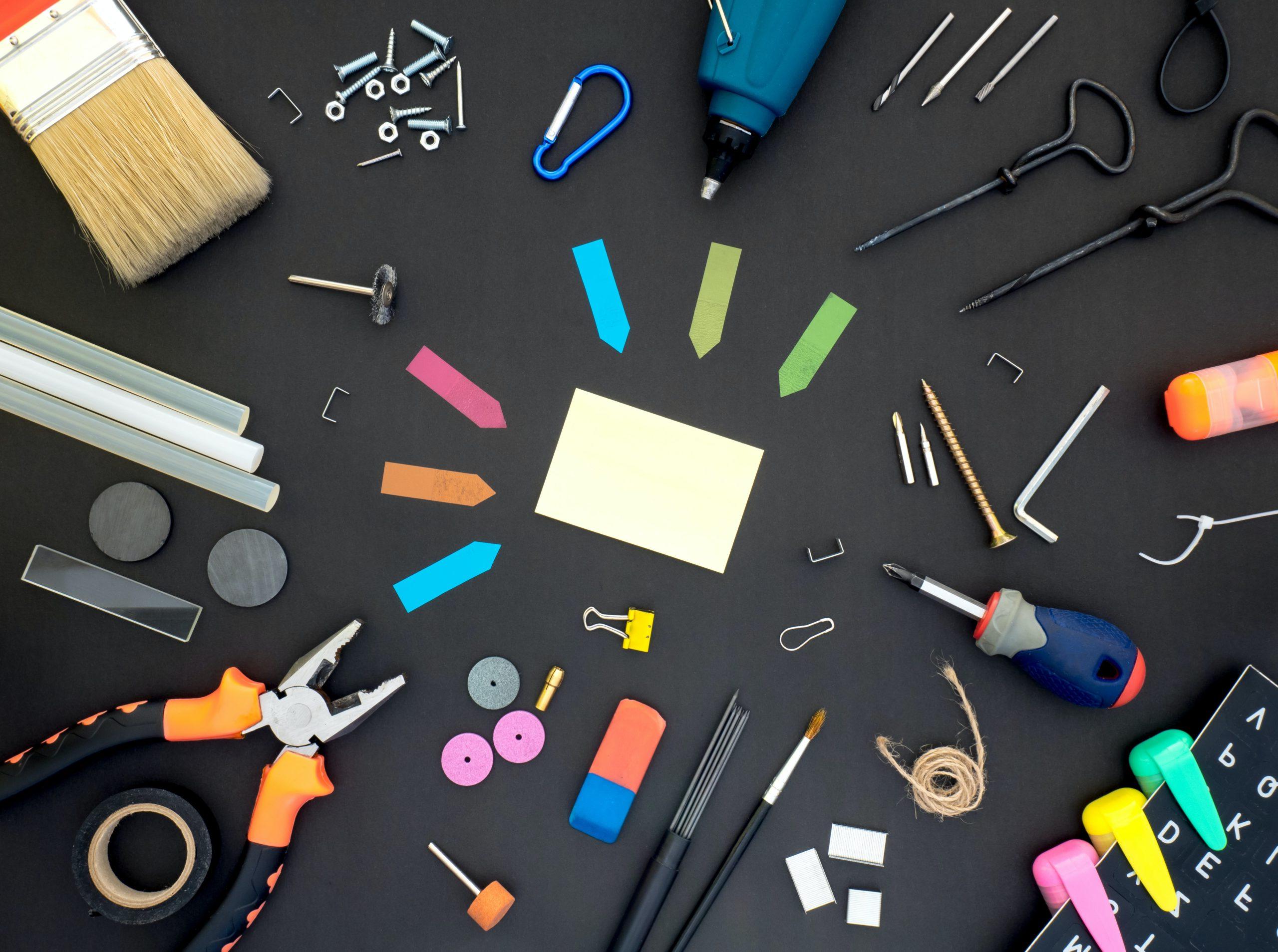 各种各样的工具放在黑色的表面上，包括便利贴, screwdrivers, wrenches, tape, paintbrushes, highlighters, glue sticks, staples, magnets, and paperclips.