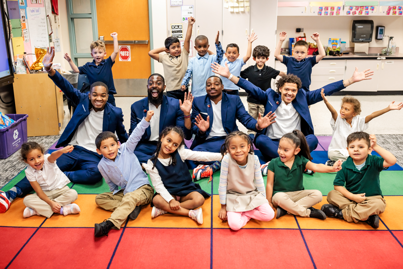的 密歇根州立大学丹佛 Call Me MISTER students and director sit with a group of elementary school students on a colorful floor mat, arms extended and smiling.